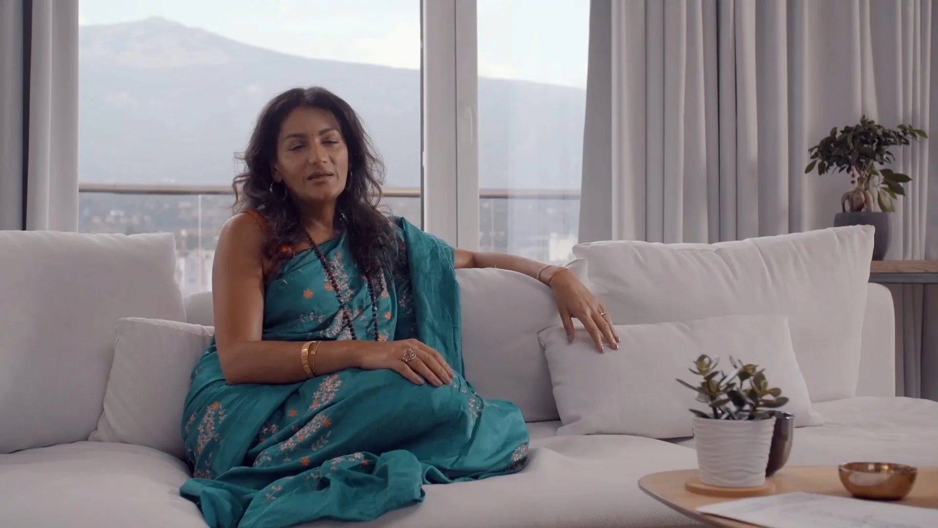 A woman in a teal sari resting on a white sofa, mountains through the window behind her